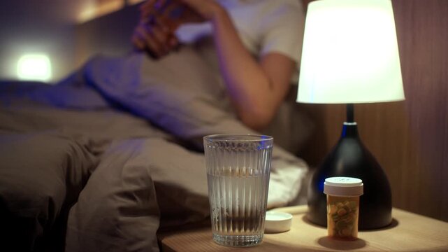Woman taking sleeping pills before bedtime while sitting on bed at home. Sleep aid medication, nighttime routine, stress management, healthcare and wellness concept.
