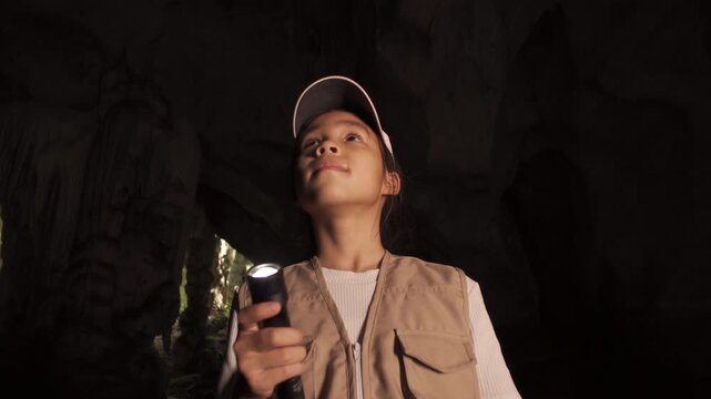 Happy little Asian girl explorer holding a flashlight and looking up inside a dark cave.