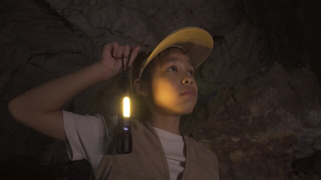 Little Asian girl explorer holding up a flashlight to illuminate a dark underground cave.