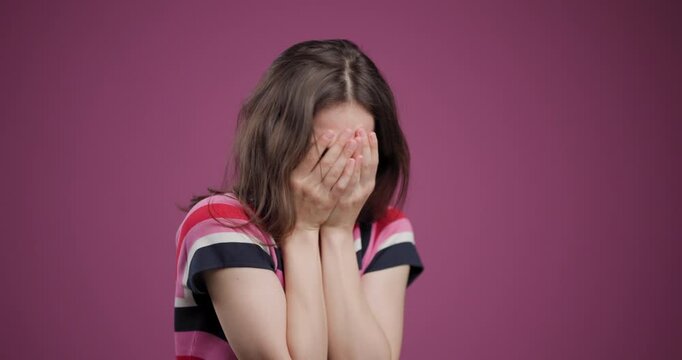 Young woman covering her face with hands against a pink background, expressing sadness or frustration