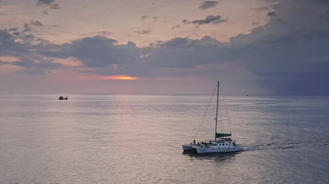 Catamaran with passengers is sailing on calm Andaman Sea waters during beautiful Phuket sunset, with small fishing boats visible in the distance and a colorful sky. Evening boat tour, summer vacation