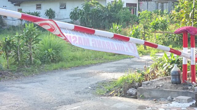 Red and white striped boom barrier gate with hanging banner blocking narrow village road surrounded by green plants