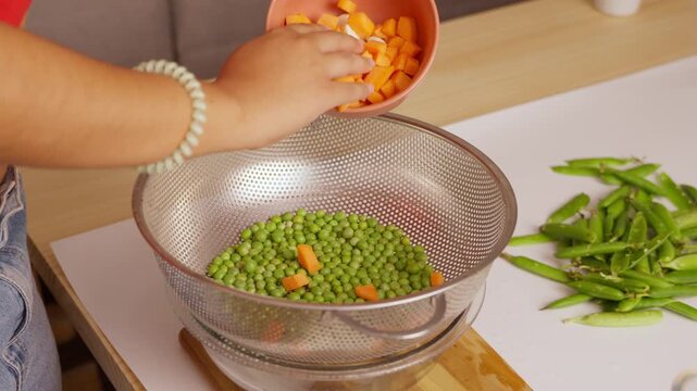 Person pouring diced carrots into colander with green peas in slow motion