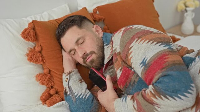 Man holding smartphone and tapping screen while nudging sleeping partner on bed with orange pillow and guitar in building; quiet frustration.