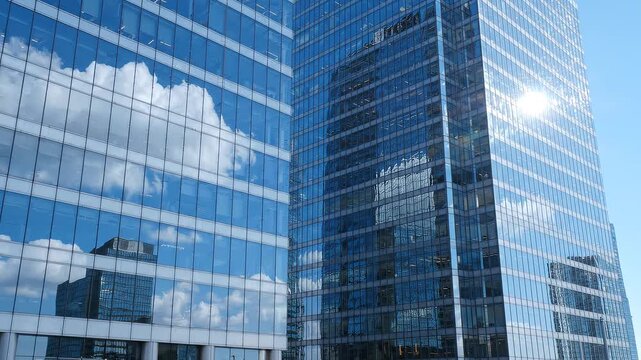 Glass skyscraper facade reflecting white clouds and blue sky with sun glare