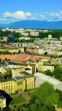 Vertical video. Geneva, Switzerland. Aerial flight over Plaine de Plainpalais, its distinctive red diamond-shaped esplanade and surrounding historic cityscape framed by the Jura Mountains under a bri