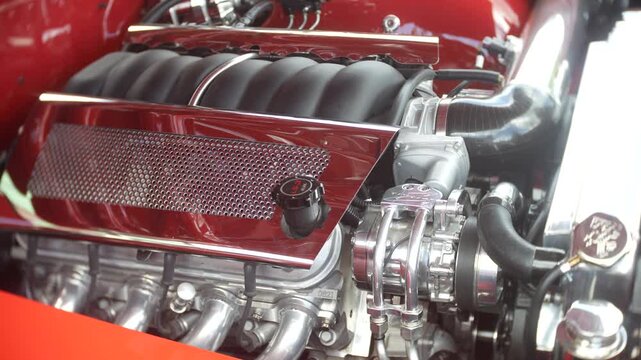 Engine exposed inside a custom hot rod shows polished LS3 components and intake detail. Close-up view highlights hoses, belts, metal mesh cover, and orange bodywork