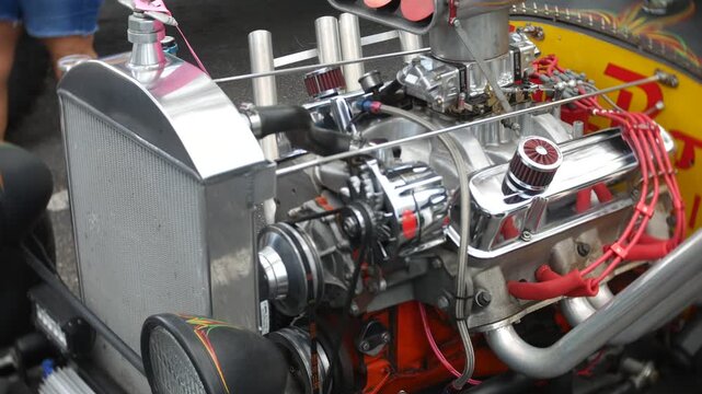 Engine parts exposed on a custom hot rod at a car show. Close-up view shows chrome radiator, belts, headlights, racing tires, and polished mechanical detail