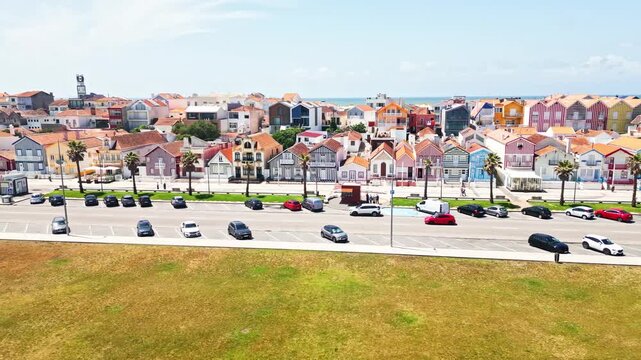Drone panning left across colourful striped houses and coastal road in Aveiro, Portugal, showing seaside architecture, vehicles and city surroundings