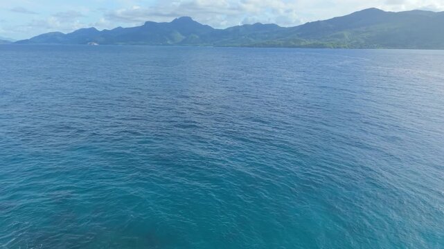 Drone pullback reveal shot showing a tiny rocky islet with palm trees surrounded by waves, with the majestic mountains of Mahe island in the background at Anse Soleil.