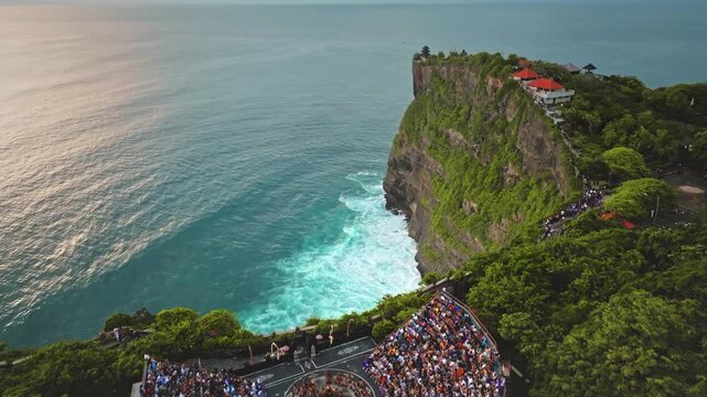 Aerial Uluwatu Temple, perched on a cliff edge, features a traditional Balinese Kecak dance in an outdoor amphitheater filled with spectators, offering stunning views of the Indian Ocean during sunset