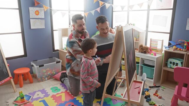 Boy drawing on easel whiteboard with marker while two men watch and guide in a brightly furnished playroom; playful learning joy.