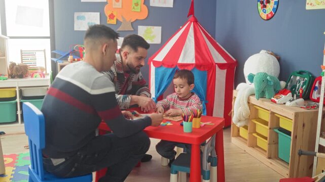 Man guides boy's hand while two adults help arrange colorful tokens, crayons and markers on a small red play table in a playroom inside a building; playful bonding.