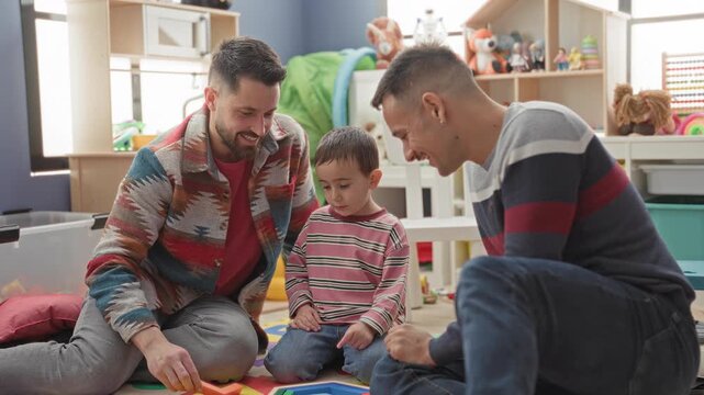 Two men guide little boy assembling colorful wooden blocks on foam playmat inside building playroom, surrounded by toys and shelves; gentle bonding joy.
