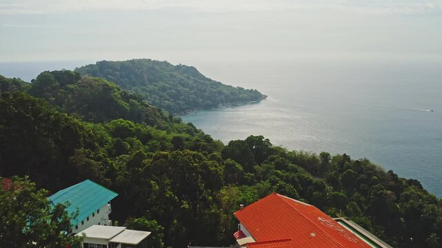 Modern white villa with an orange roof nestled in lush green tropical trees, offering panoramic views of the Andaman Sea and the coastline of Phuket, Thailand, with a boat cruising in the distance