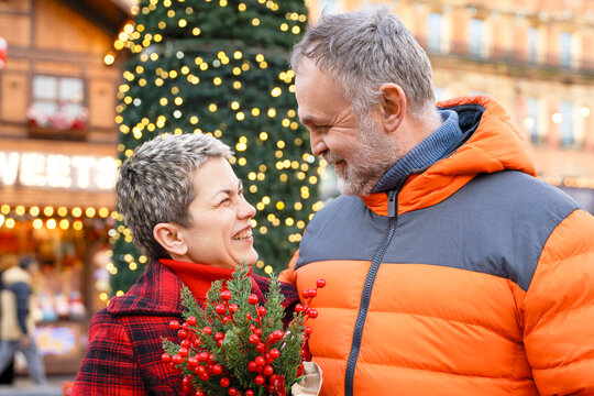 Couple smiles at each other while holding holiday decorations in a festive market during winter