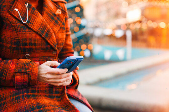 Person sits outdoors in a checked coat using smartphone while lights shine in the background near a water feature in the evening