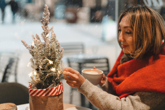 Woman enjoys warm drink while sitting near decorated small tree in city cafe during winter season