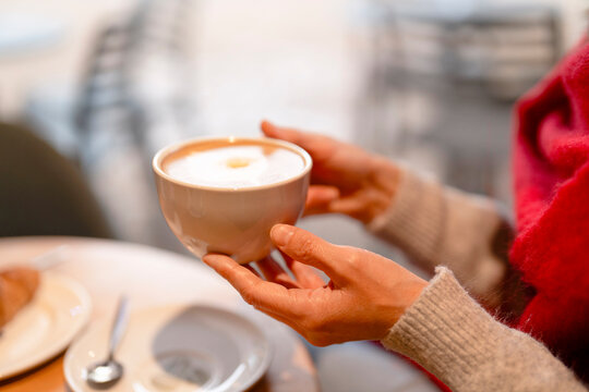 Coffee cup held by a person in a cozy cafe setting during afternoon hours with a croissant on the table nearby