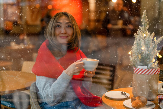 Woman enjoying hot drink in cafe while snow falls outside during winter season