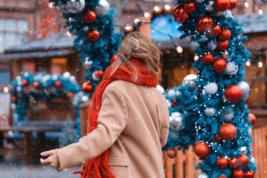 Woman walks through a decorated street with snow falling and colorful ornaments on a winter day in a festive city scene