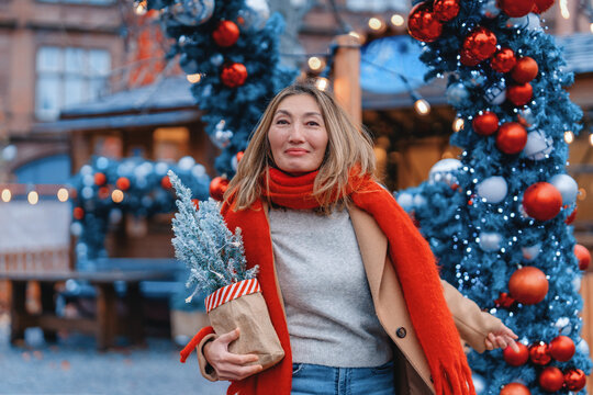 Woman enjoying festive decorations at a market while holding a small Christmas tree in a bag on a winter day
