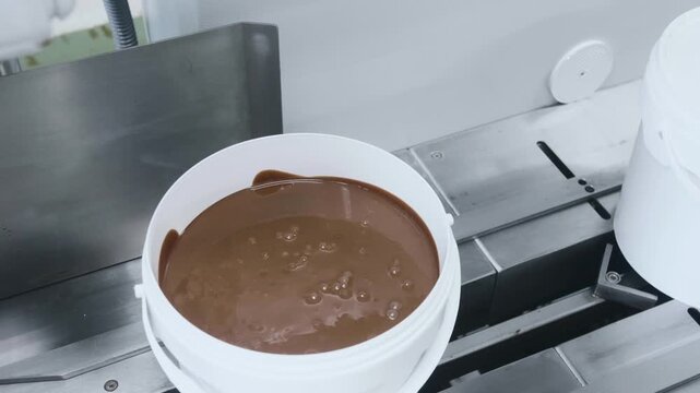 A close-up of creamy chocolate mixture inside a plastic container moving along an industrial conveyor system during automated food processing and packaging operations