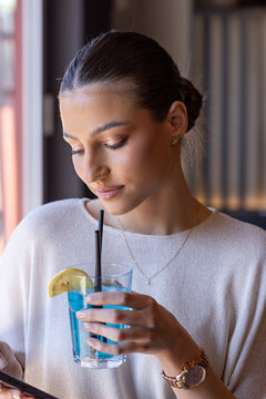 Gentle portrait of a young woman holding a blue cocktail and looking down at her phone