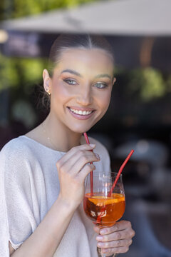 Smiling young woman with an orange cocktail shot through a glass window with soft reflections