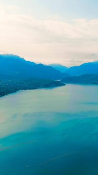 Vertical video. Annecy, France. Lake Annecy, turquoise glacial waters framed by layered Alpine peaks and lakeside villages, aerial panoramic view in summer morning light. Drone footage, Departure of 