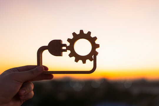 Hand holding wooden gear and plug symbol against a golden sunset sky. Concept of renewable energy, green technology, industrial innovation, and sustainable power solutions for a clean future.