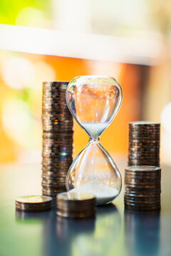 Hourglass with flowing sand surrounded by stacked coins on a desk. Concept of time is money, financial investment, business growth, wealth management, retirement planning, and savings.