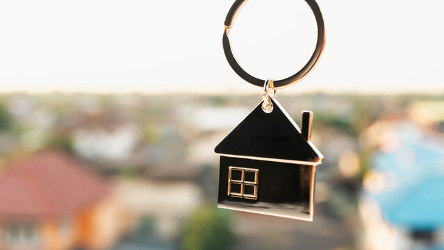 Close up of a metal house shape keychain against a blurred residential neighborhood background at sunset. Concept of real estate, property investment, buying a new home, and mortgages.