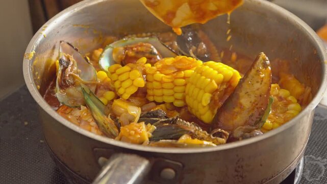 A person stirring a spicy seafood boil platter featuring mussels, shrimp, shellfish, sweet corn, and a rich spicy savory sauce