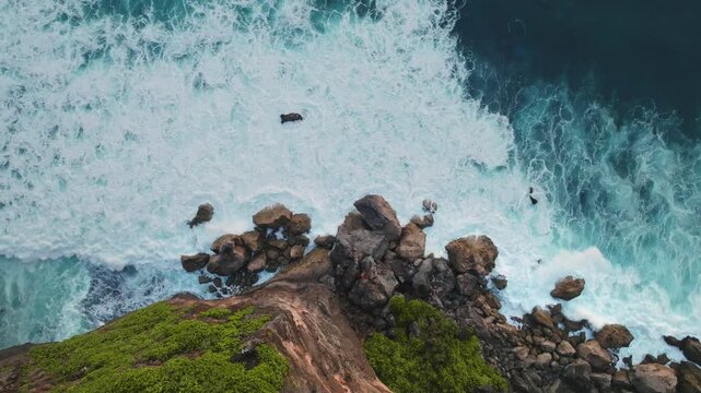 Crashing ocean waves create white foam against the rugged cliffside and rocky beach below, highlighting the natural beauty and power of the sea in Bali, Indonesia. Aerial view tod down shot