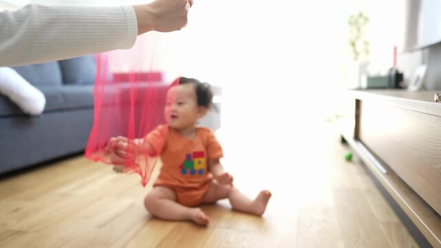 Soft light fills cozy living room space. Baby sits barefoot on warm wood floor. Adult gently holds pink sheer fabric. Child reaches out curiously, eyes bright. Playful interaction sparks joy