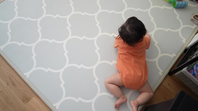 Tiny toddler crawls on a stylish gray mat. Child wears an orange one-piece outfit. Baby moves slowly, focused on toys nearby. Mat features elegant white geometric pattern