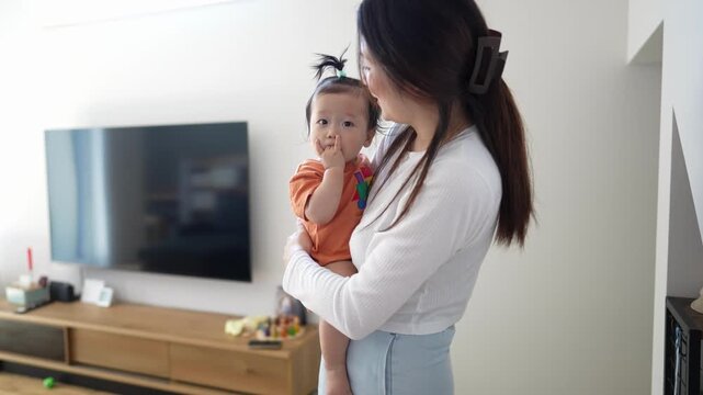 Mother holds baby gently, smiling softly. Baby looks curious, reaching for own mouth. Living room background has TV and wooden furniture. Ideal for parenting, family, or daily life content