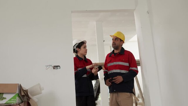 Two construction workers wearing safety helmets discussing renovation progress and reviewing plans inside unfinished building interior.