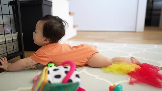 Baby crawls toward black object with curious interest. Child wears orange top, barefoot, focused on object. Soft rug, scattered toys, bright colors fill scene