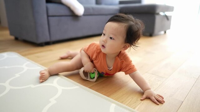 Toddler crawls on wooden floor with green ball. Child wears orange outfit, moves slowly. Soft light highlights baby's curious expression. Mat with white pattern lies beneath