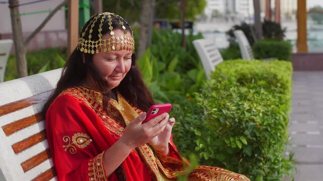 Woman in red traditional dress with gold zari embroidery, ornate gold headpiece sits on bench, using pink smartphone in outdoor garden setting with greenery.