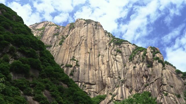 A crisp low-angle panning shot looking up at a massive, vertical granite rock face on Mount Hua under a bright blue sky filled with moving white puffy clouds.