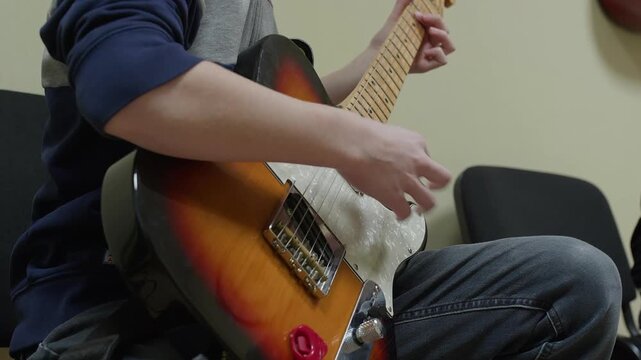 Student musician playing sunburst electric guitar while seated, demonstrating finger placement on fretboard during practice or lesson.