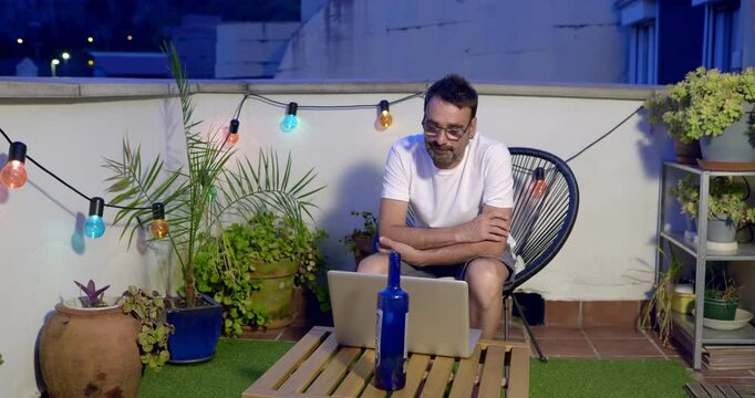 Remote Work Oasis: A man relaxes on a rooftop patio, illuminated by string lights, as he works on his laptop amidst lush greenery and a serene evening ambiance.