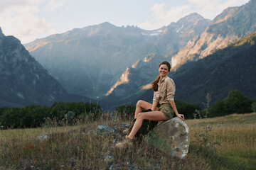 Obraz premium Young woman sitting on rock in mountain meadow, smiling relaxed, casual outfit, sunny day, natural landscape, outdoor lifestyle, scenic background