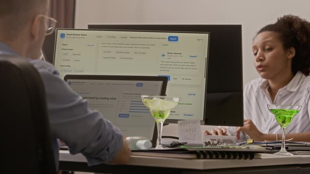 Two biracial colleagues sitting at desks, working on computers and laptop in stuffy office during summer day