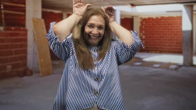 Woman raising hands to head making playful horns while wearing a striped blouse in a building; playful joy.