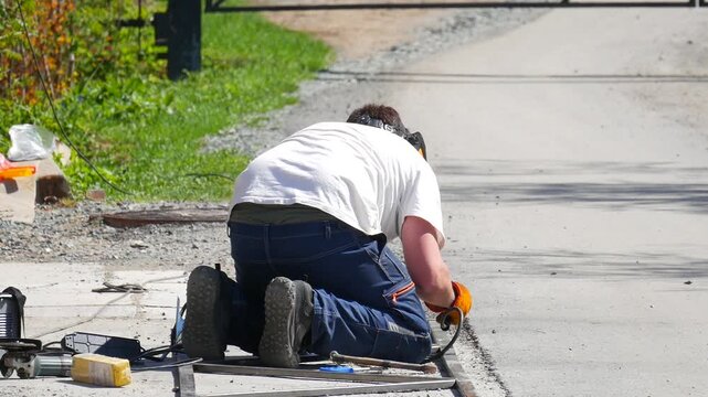 Welder man kneeling to repair metal gate on outdoor road. Laborer working with grinding tool to restore hardware. Industrial maintenance and construction activity.