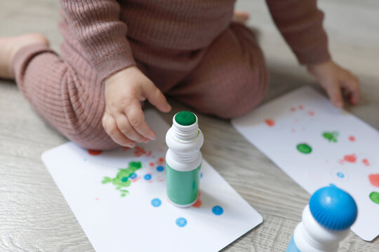 Close up on toddler hands playing with green bingo dauber marker on floor. Little child drawing colorful paint dots on white paper sheets during home activity. Fine motor skills development and